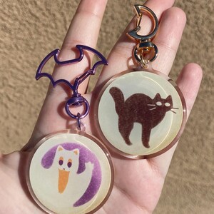 May include: Two round, clear acrylic keychains with a gold metal clasp. One keychain features a white cat with a purple tail and the other keychain features a brown cat. Both keychains have a vintage, faded look.