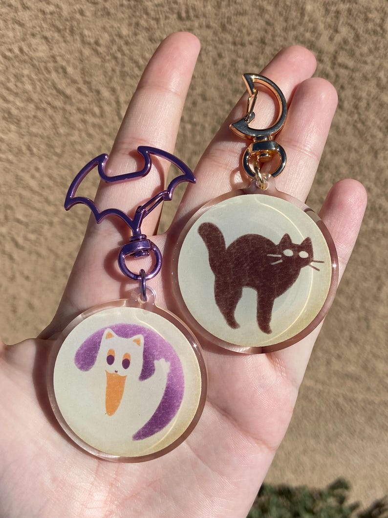 May include: Two round, clear acrylic keychains with a gold metal clasp. One keychain features a white cat with a purple tail and the other keychain features a brown cat. Both keychains have a vintage, faded look.