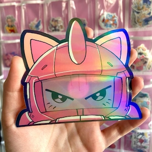 May include: A pink holographic sticker featuring a cartoon character with cat ears and an angry expression. The sticker has a helmet-like design with a visor and is held in a hand.