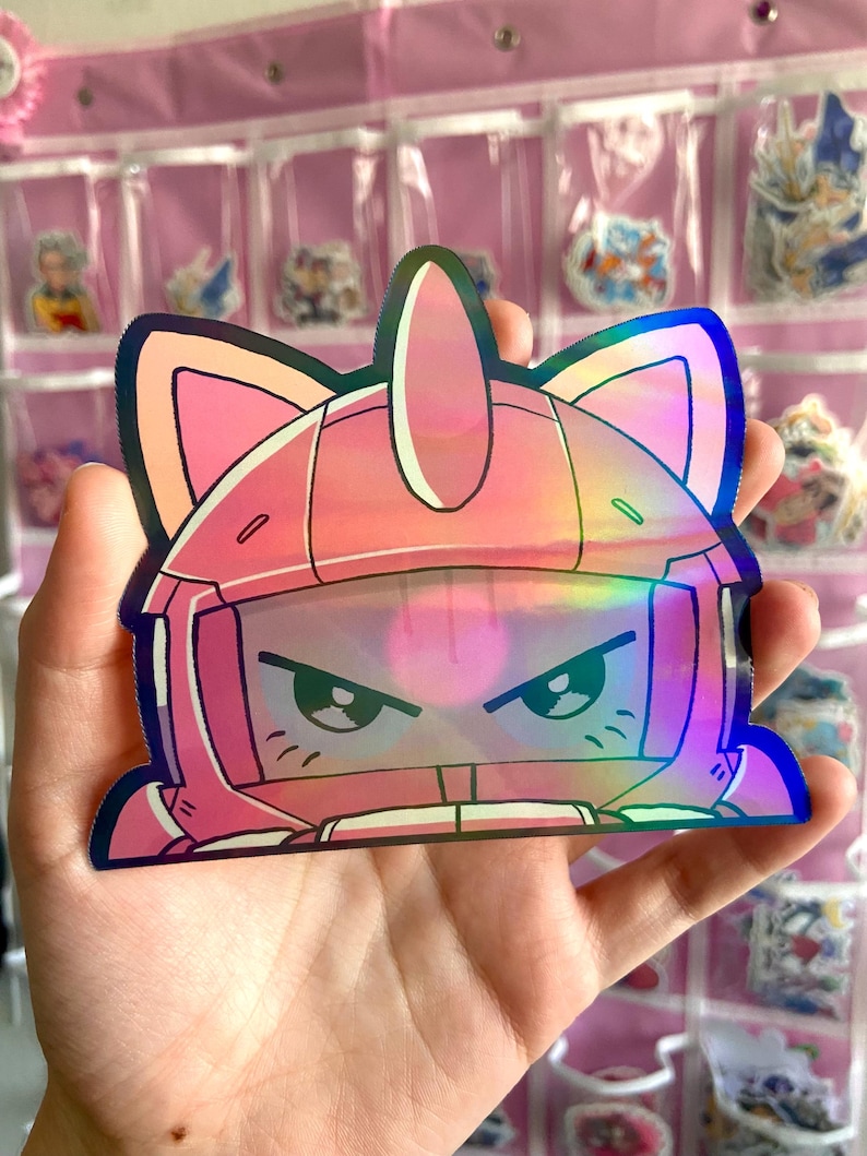 May include: A pink holographic sticker featuring a cartoon character with cat ears and an angry expression. The sticker has a helmet-like design with a visor and is held in a hand.