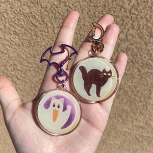 May include: Two round, clear, resin keychains with a purple and gold metal clasp. One keychain features a white ghost with orange eyes and a purple background. The other keychain features a black cat with orange eyes and a white background.