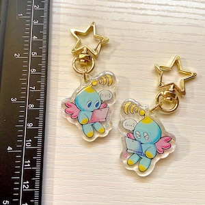 May include: Two clear acrylic charms featuring a blue and yellow cartoon character with wings holding a laptop. The charms are attached to gold star-shaped keychains.