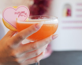 May include: A pink heart-shaped cocktail stirrer with the text "FUTURE Mrs. B" on it, placed in a glass of orange juice. The glass is being held by a hand.
