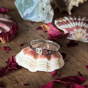 May include: A silver ring featuring a crescent moon and full moon design. The ring is displayed on a seashell, surrounded by red rose petals and other seashells. The ring is a piece of jewelry.