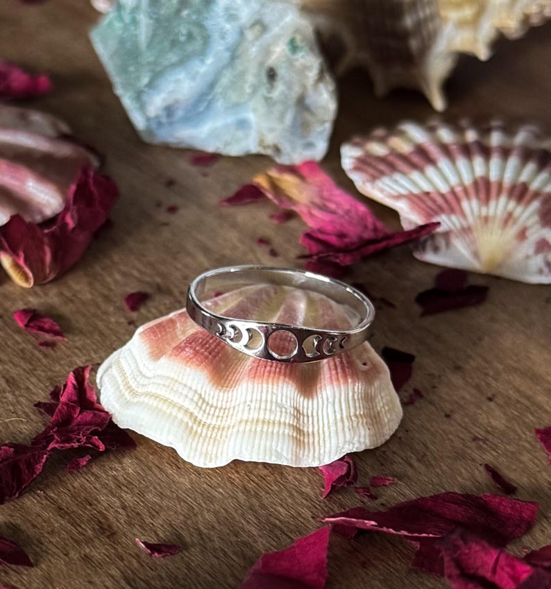 May include: A silver ring featuring a crescent moon and full moon design. The ring is displayed on a seashell, surrounded by red rose petals and other seashells. The ring is a piece of jewelry.