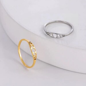 May include: Two rings, one gold and one silver, each featuring a crescent moon design. The gold ring is in focus, showcasing a delicate, openwork pattern of moon phases. The silver ring is in the background, also displaying a similar design.