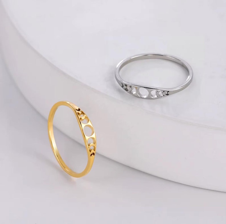 May include: Two rings, one gold and one silver, each featuring a crescent moon design. The gold ring is in focus, showcasing a delicate, openwork pattern of moon phases. The silver ring is in the background, also displaying a similar design.