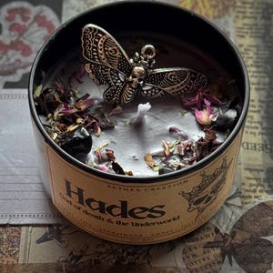 以下が含まれることがあります： 「Hades God of death & the Underworld」の文字が入った丸い缶に入った黒いキャンドル。キャンドルには、銀色の蝶のチャーム、ドライフラワー、小さな黒い石が付いています。サンダルウッドとアンバーの香りです。