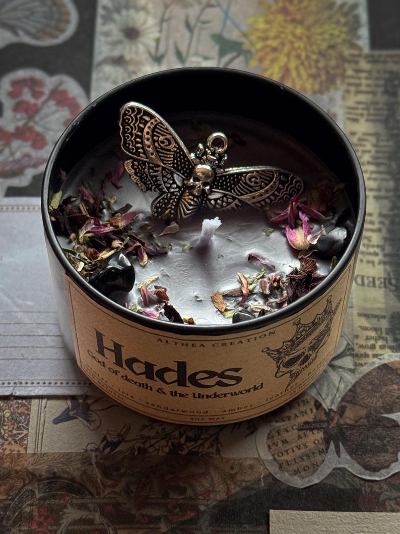以下が含まれることがあります： 「Hades God of death & the Underworld」の文字が入った丸い缶に入った黒いキャンドル。キャンドルには、銀色の蝶のチャーム、ドライフラワー、小さな黒い石が付いています。サンダルウッドとアンバーの香りです。
