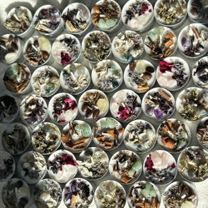May include: Assortment of small, white tea light candles with various natural ingredients and crystals embedded in the wax. The candles are arranged in a grid pattern.