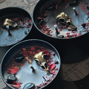 May include: Three black candles in round metal containers, each featuring a central wick, a metallic gold-toned bird skull charm, scattered dried red flower petals, and small polished black stones embedded in the black wax surface.