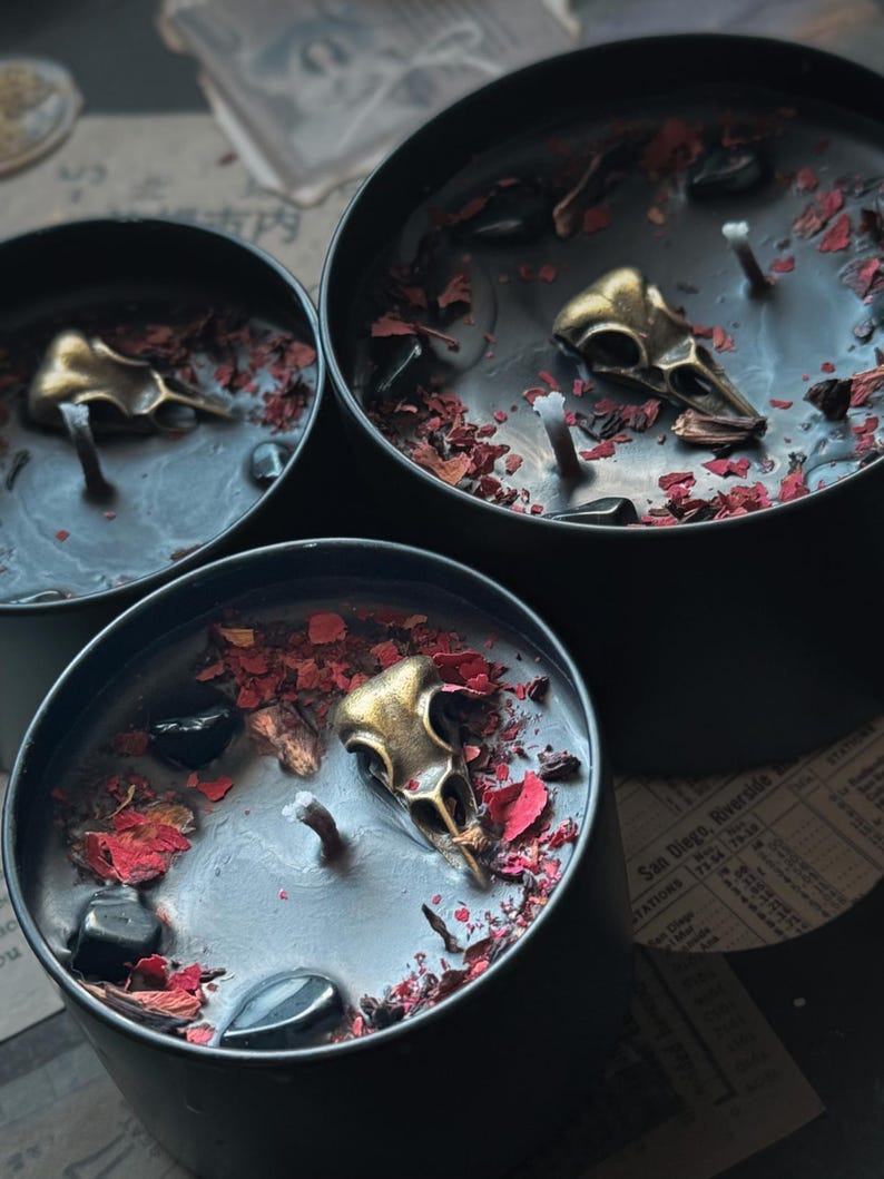 May include: Three black candles in round metal containers, each featuring a central wick, a metallic gold-toned bird skull charm, scattered dried red flower petals, and small polished black stones embedded in the black wax surface.