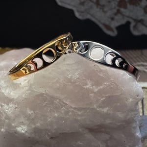 May include: Two crescent moon phase bangles. One is gold-toned, the other is silver-toned. Each bangle features cut-out moon phases. The bangles are displayed on a light pink, textured surface.
