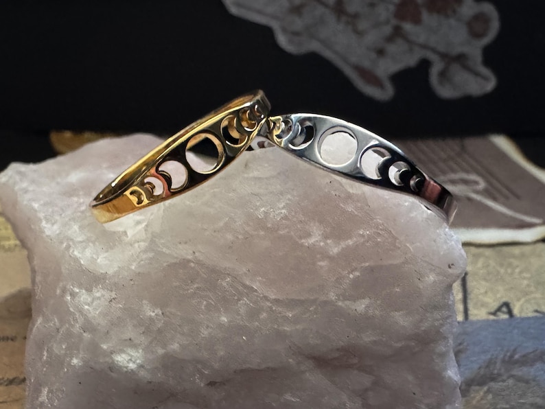 May include: Two crescent moon phase bangles. One is gold-toned, the other is silver-toned. Each bangle features cut-out moon phases. The bangles are displayed on a light pink, textured surface.