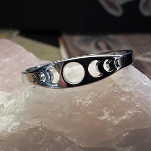 May include: A silver ring featuring a series of crescent moon cutouts. The ring is set against a light pink, textured surface, with a blurred background. The ring's design showcases the phases of the moon.