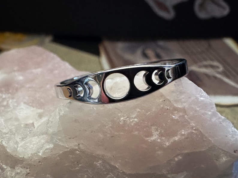 May include: A silver ring featuring a series of crescent moon cutouts. The ring is set against a light pink, textured surface, with a blurred background. The ring's design showcases the phases of the moon.