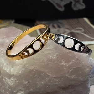 May include: A gold-colored ring and a silver-colored bracelet, both featuring a crescent moon design. The ring is oval-shaped with cut-out crescent moon shapes. The bracelet is a thin band with similar cut-out moon shapes. Both are displayed on a light pink stone.
