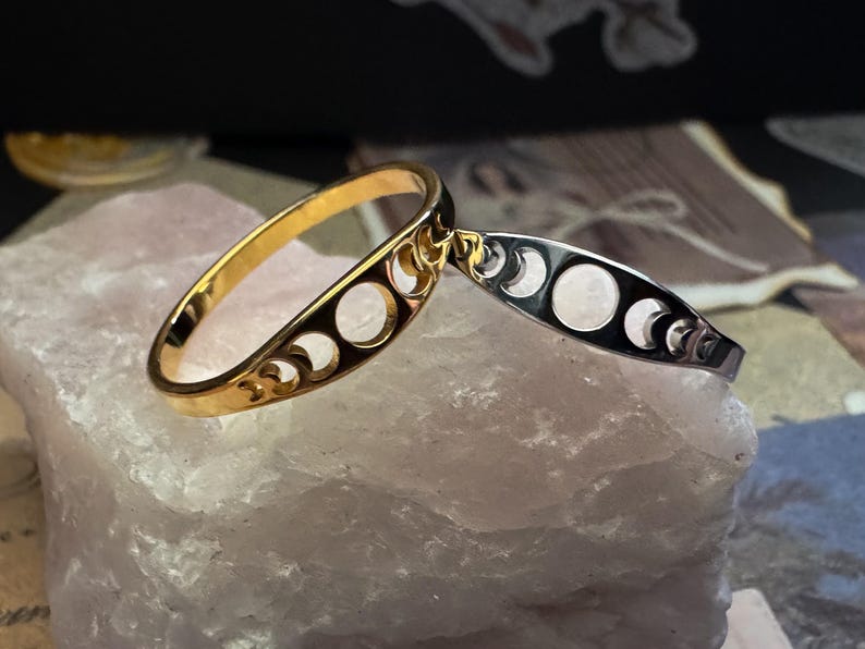 May include: A gold-colored ring and a silver-colored bracelet, both featuring a crescent moon design. The ring is oval-shaped with cut-out crescent moon shapes. The bracelet is a thin band with similar cut-out moon shapes. Both are displayed on a light pink stone.