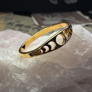 May include: A gold-colored ring with a crescent moon and circle design. The ring is set on a white, textured surface. The ring's design features a series of crescent moons and circular cutouts, creating a celestial theme. The ring is a jewelry item.