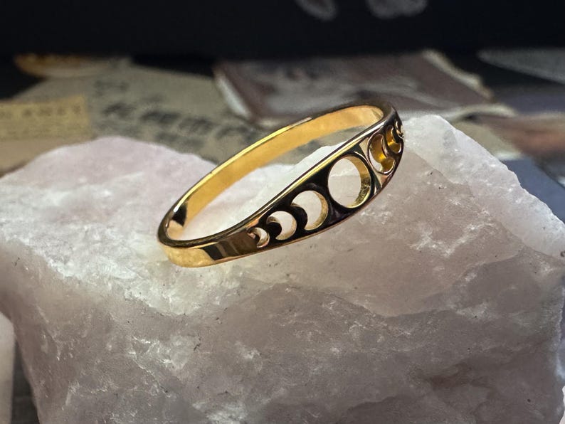 May include: A gold-colored ring with a crescent moon and circle design. The ring is set on a white, textured surface. The ring's design features a series of crescent moons and circular cutouts, creating a celestial theme. The ring is a jewelry item.