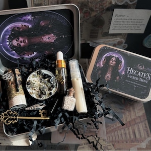 May include: A metal gift tin labeled Hecate's Sacred Trove containing a candle with herbs, a glass dropper bottle with oil, a spray bottle with cream, a small corked vial of sand, a gold-toned skeleton key, and a seashell nestled in black paper.