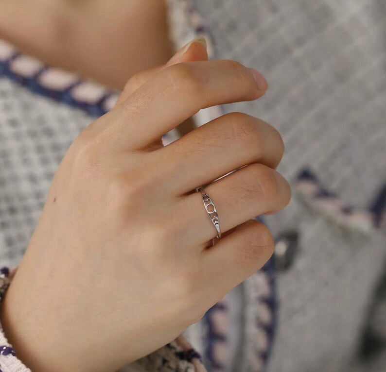 May include: A delicate silver ring with an openwork design featuring a central circle and decorative accents. The ring is worn on a finger, showcasing its intricate details and subtle elegance. The ring's design is simple yet sophisticated.