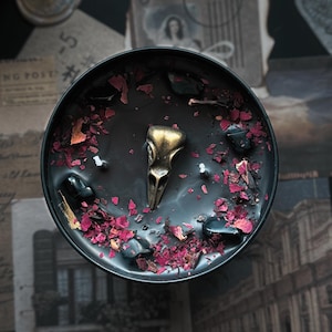 May include: A round black candle in a container featuring a central metallic gold bird skull ornament, two white wicks, scattered dark polished stones, and dried pink and red flower petals embedded in the smooth black wax surface.
