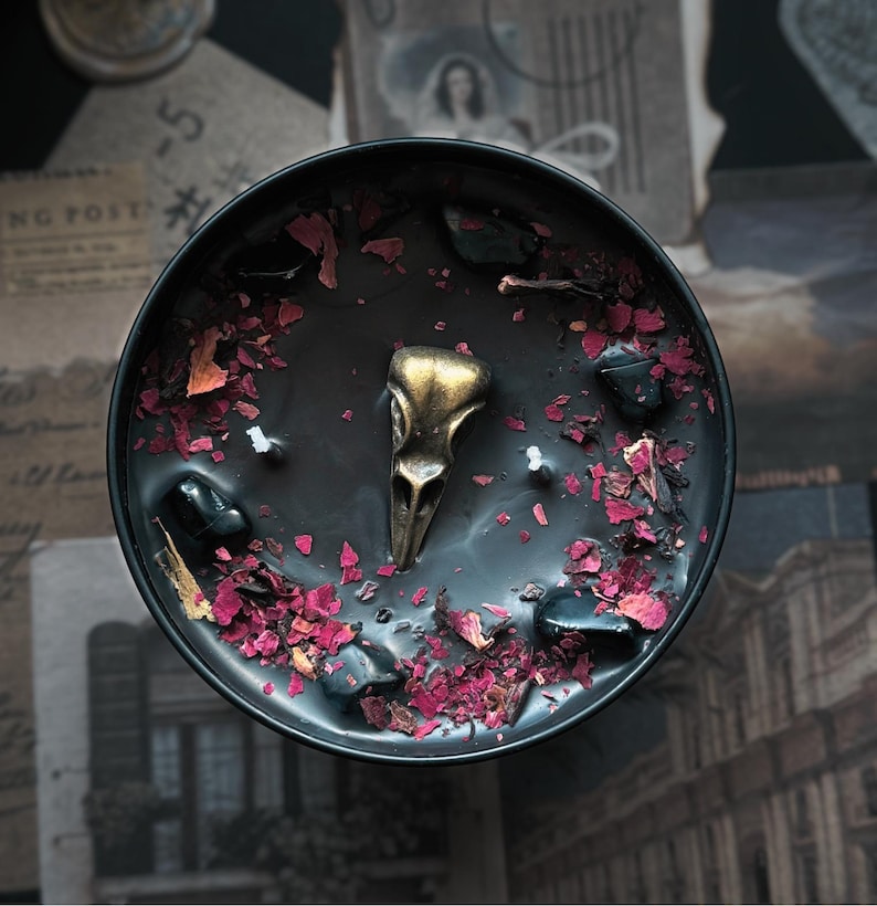 May include: A round black candle in a container featuring a central metallic gold bird skull ornament, two white wicks, scattered dark polished stones, and dried pink and red flower petals embedded in the smooth black wax surface.