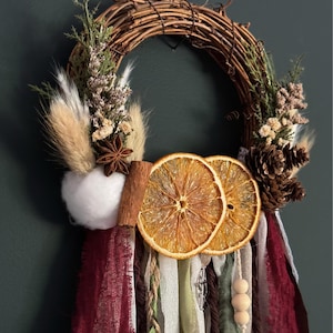 Puede incluir: Una corona de sarmientos decorada con rodajas de naranja seca, una rama de canela, piñas, bolas de algodón blanco y hierbas secas. Largas tiras de cintas de tela en color burdeos, verde salvia y crema cuelgan de la parte inferior.