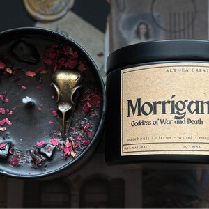 May include: A black soy wax candle in a metal tin with a label reading Morrigan Goddess of War and Death. The candle surface features black wax, scattered pink flower petals, dark stones, and a metallic gold-toned bird skull charm.