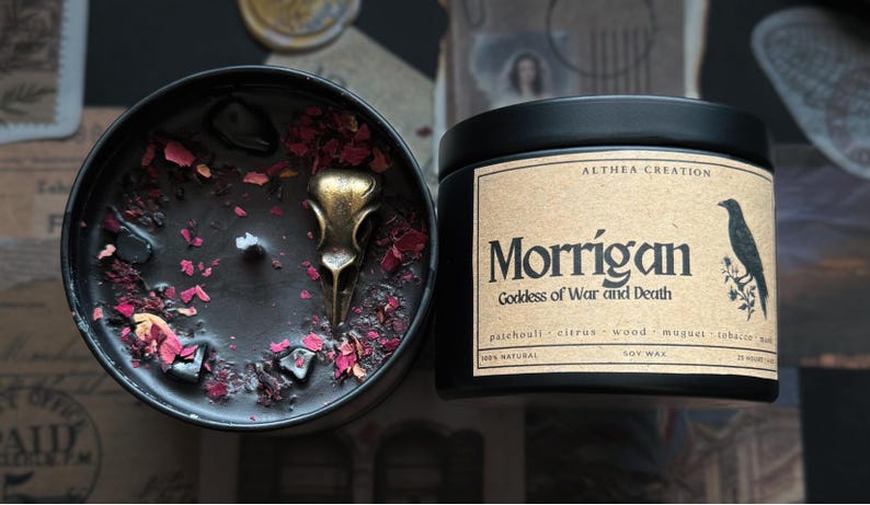 May include: A black soy wax candle in a metal tin with a label reading Morrigan Goddess of War and Death. The candle surface features black wax, scattered pink flower petals, dark stones, and a metallic gold-toned bird skull charm.