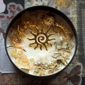 Op de afbeelding: Een ronde zwarte kaars met een gouden zonvormige bedel in het midden. De kaars is omgeven door gedroogde bloemen, kruiden en kleine gele kristallen. De kaars zit in een zwarte container.