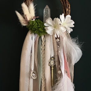 May include: A wall hanging featuring a circular grapevine wreath adorned with a clear quartz crystal, white flowers, green moss, and dried grasses. Long fabric ribbons in beige, sage, and white hang down, accented with a brass key, a metal charm, and white feathers.