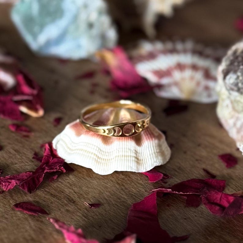 May include: A gold-colored ring with a crescent moon design, resting on a seashell. The ring is the focal point, with the seashell and scattered rose petals adding to the aesthetic. The ring's design features three crescent moons.