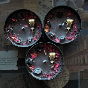 May include: Three round black candles in metal tins, each featuring a central brass-colored bird skull charm, scattered dark polished stones, and dried red flower petals embedded in the black wax surface, viewed from directly above.