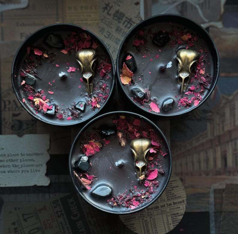 May include: Three round black candles in metal tins, each featuring a central brass-colored bird skull charm, scattered dark polished stones, and dried red flower petals embedded in the black wax surface, viewed from directly above.