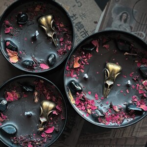 May include: Three black round candles in metal tins, each featuring a central gold-toned bird skull ornament, scattered black polished stones, and dried pink and red flower petals embedded in the dark wax surface around a single white wick.
