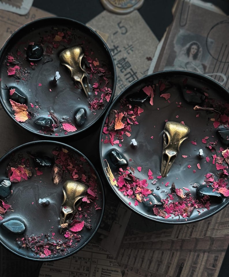 May include: Three black round candles in metal tins, each featuring a central gold-toned bird skull ornament, scattered black polished stones, and dried pink and red flower petals embedded in the dark wax surface around a single white wick.