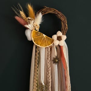 Puede incluir: Una corona circular de vid adornada con una rodaja de naranja seca, una flor de ganchillo y hierbas secas. Cuelgan largas cintas de tela en tonos beige, crema y óxido, con un pequeño dije de sol de metal y una llave de estilo antiguo.