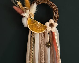 May include: A circular grapevine wreath adorned with a dried orange slice, a crocheted flower, and dried grasses. Long, hanging fabric ribbons in shades of beige, cream, and rust are attached, featuring a small metal sun charm and a vintage-style key.