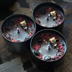 May include: Three black cylindrical candle tins filled with black wax. Each candle features a central wick, a metallic gold-toned bird skull ornament, scattered dark red dried flower petals, and small polished black stones embedded in the wax surface.