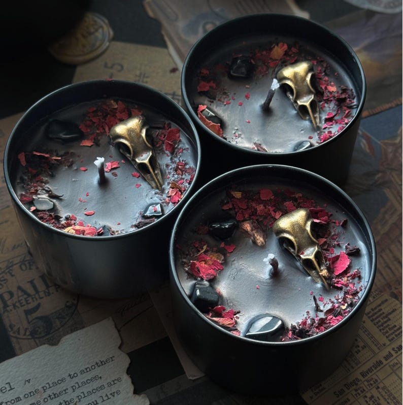 May include: Three black cylindrical candle tins filled with black wax. Each candle features a central wick, a metallic gold-toned bird skull ornament, scattered dark red dried flower petals, and small polished black stones embedded in the wax surface.
