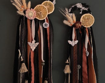 May include: Two wall hangings featuring black and white moss-covered hoops adorned with dried orange slices, wheat stalks, and flowers. Long strips of black, rust, and cream fabric hang down, accented with metal charms including a moth, pentagram, key, and bells.