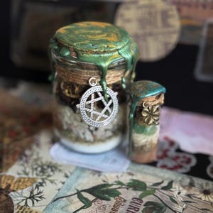 May include: Two glass jars with decorative lids. The larger jar has a green and gold wax seal, a silver pentacle charm, and is filled with layered contents. The smaller jar has a teal seal and a gold flower charm.