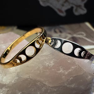 May include: Two crescent moon bangles. One is gold-toned, the other is silver-toned. Both have a series of circular cutouts and crescent moon shapes. The bangles are displayed on a light pink, textured surface.