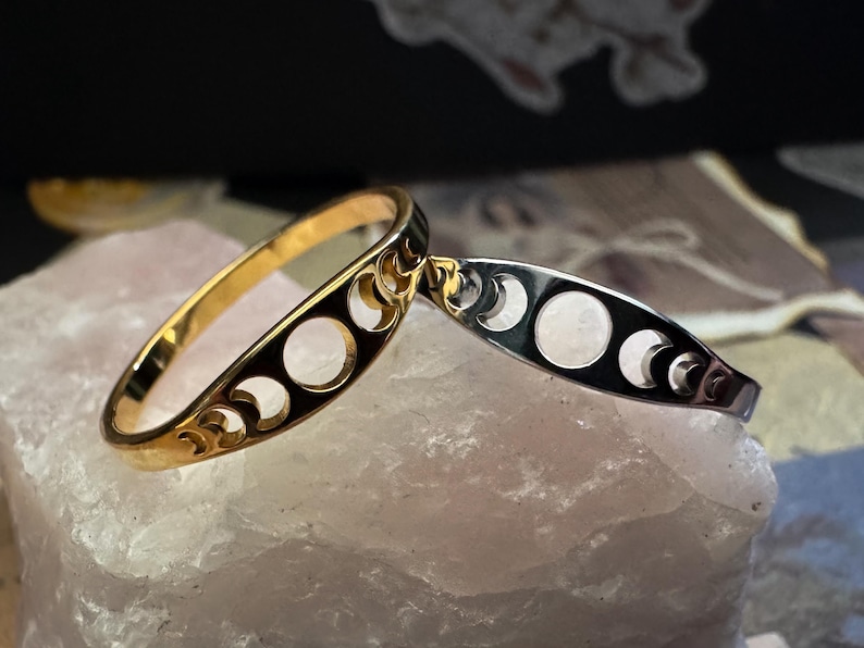 May include: Two crescent moon bangles. One is gold-toned, the other is silver-toned. Both have a series of circular cutouts and crescent moon shapes. The bangles are displayed on a light pink, textured surface.