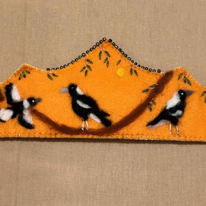 May include: An orange felt artwork depicting three magpies on a brown branch. The piece has a scalloped edge with a beaded border and a stitched outline. A small yellow sun and green leaves are also included in the design.