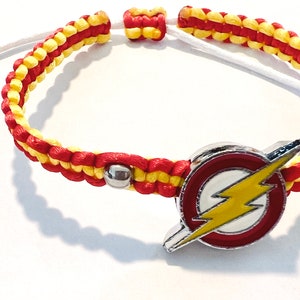 Op de afbeelding: Een rood en geel gevlochten armband met een zilveren bedel met het Flash-logo. Het logo is rood, geel en wit met een bliksem.
