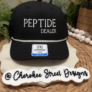 May include: A black baseball cap featuring the white embroidered text PEPTIDE DEALER on the front. The cap includes a white braided cord across the base of the crown and a rectangular Richardson Performance OSFM sticker on the center of the brim.