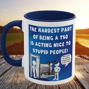 May include: White ceramic mug with a navy blue handle and interior. The mug features the text "THE HARDEST PART OF BEING A TSO IS ACTING NICE TO STUPID PEOPLE!" with a cartoon illustration of security personnel and a metal detector.